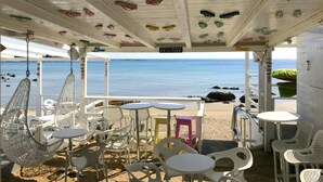 Un café frente a la playa con mobiliario blanco, una silla colgante y vistas al mar.