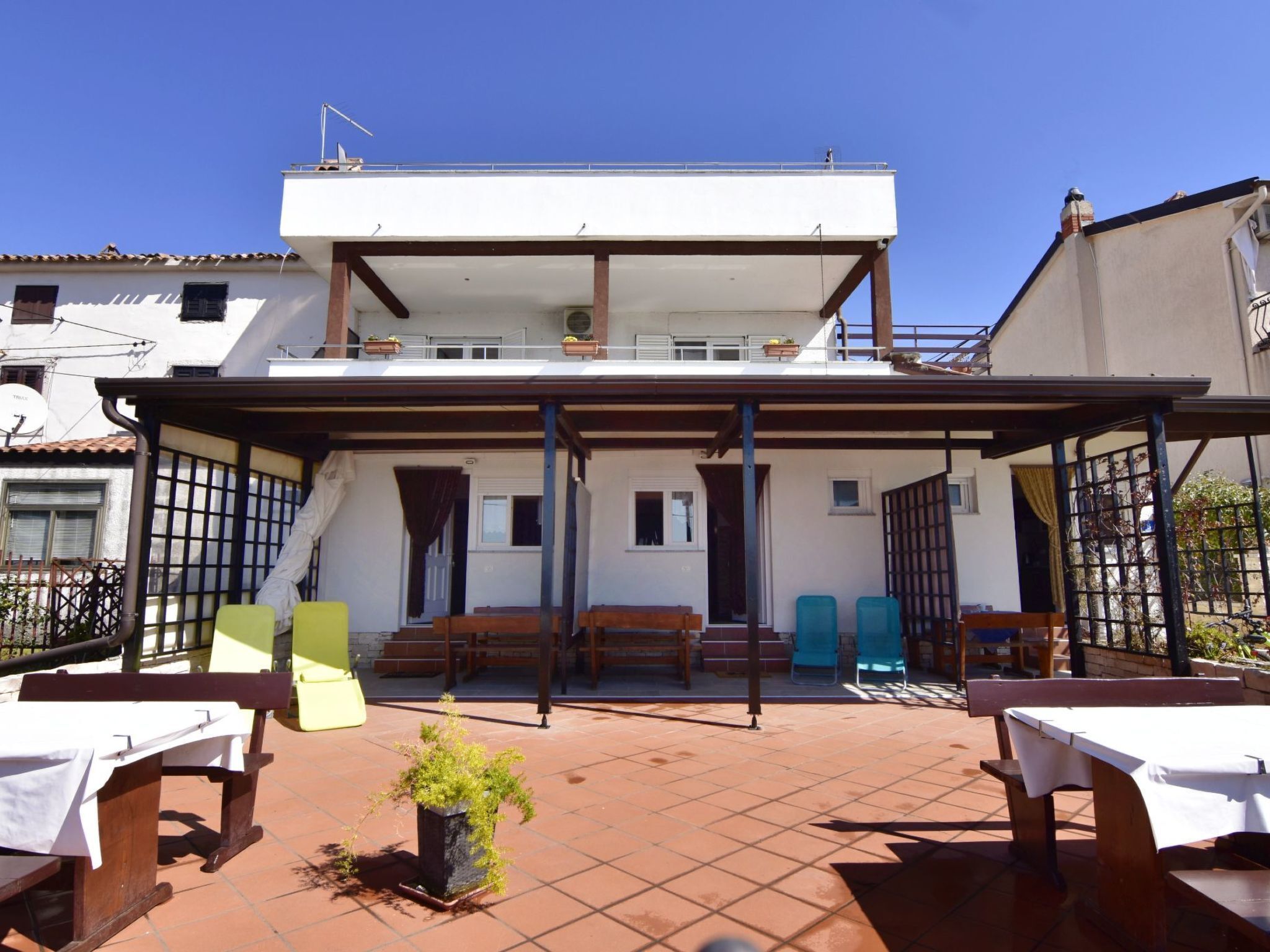 A two-story building with a covered patio area featuring wooden benches and tables, a small potted plant, and a tiled floor.