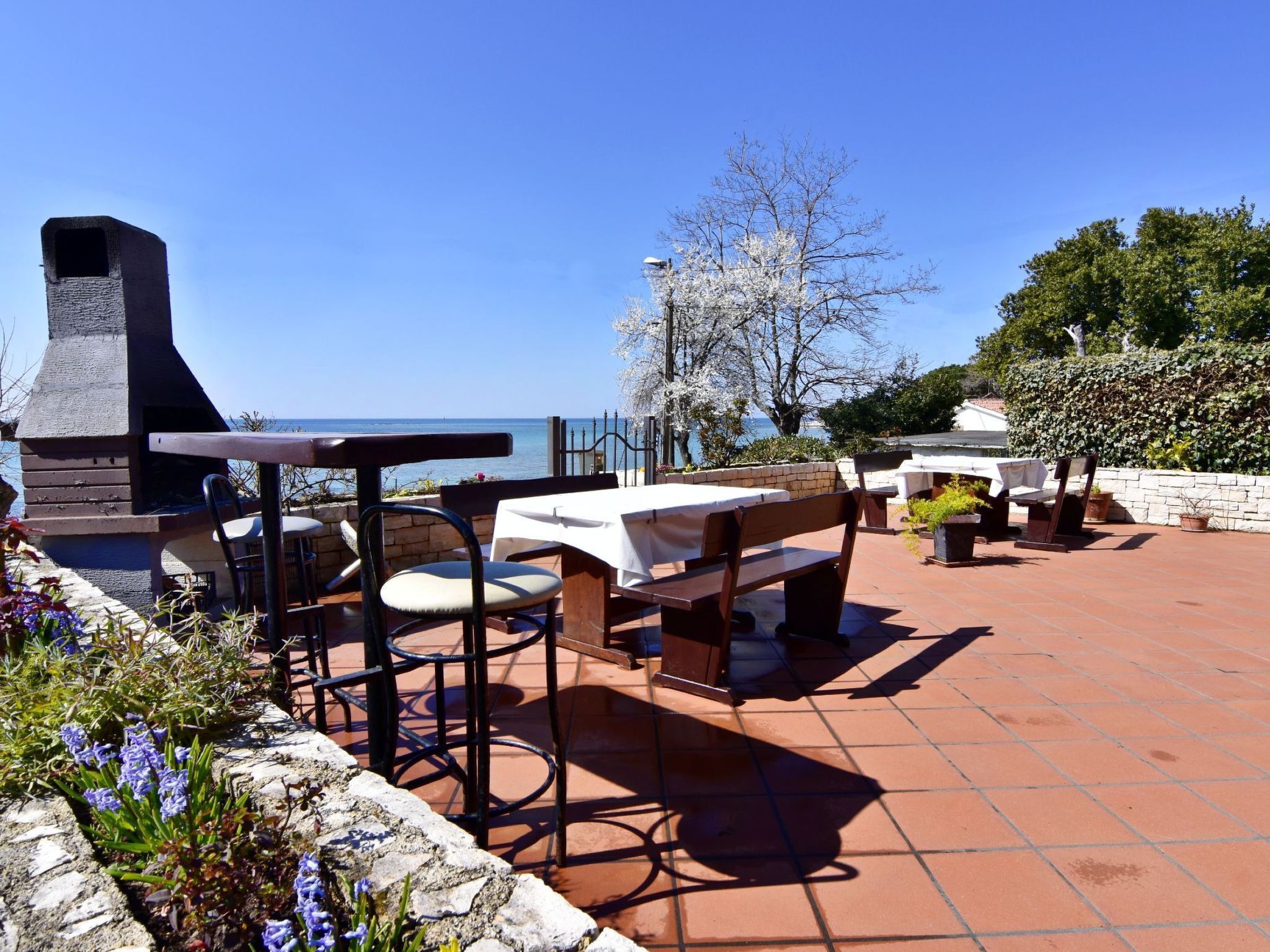 An outdoor patio with tables and chairs, a stone wall, and a view of the sea.