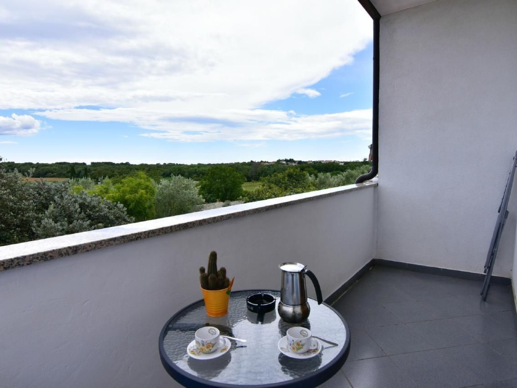 Un balcon avec une table dressée pour deux personnes, un porte-gobelet jaune, une cafetière en métal et une petite plante en pot.