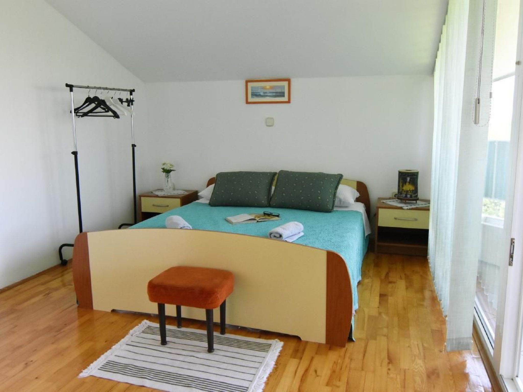 A bedroom with a bed, a nightstand, a reading lamp, a window, and a hanging clothes rack.