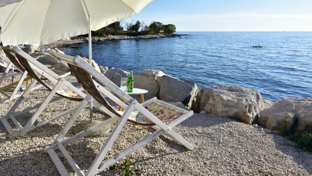 Eine Strandlage mit weißem Sonnenschirm, Holzliegen und einer felsigen Küstenlinie.