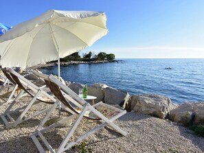A beachside setting with a white umbrella, wooden lounge chairs, and a rocky shoreline. - APP EVA by Interhome (Umag/Savudrija)