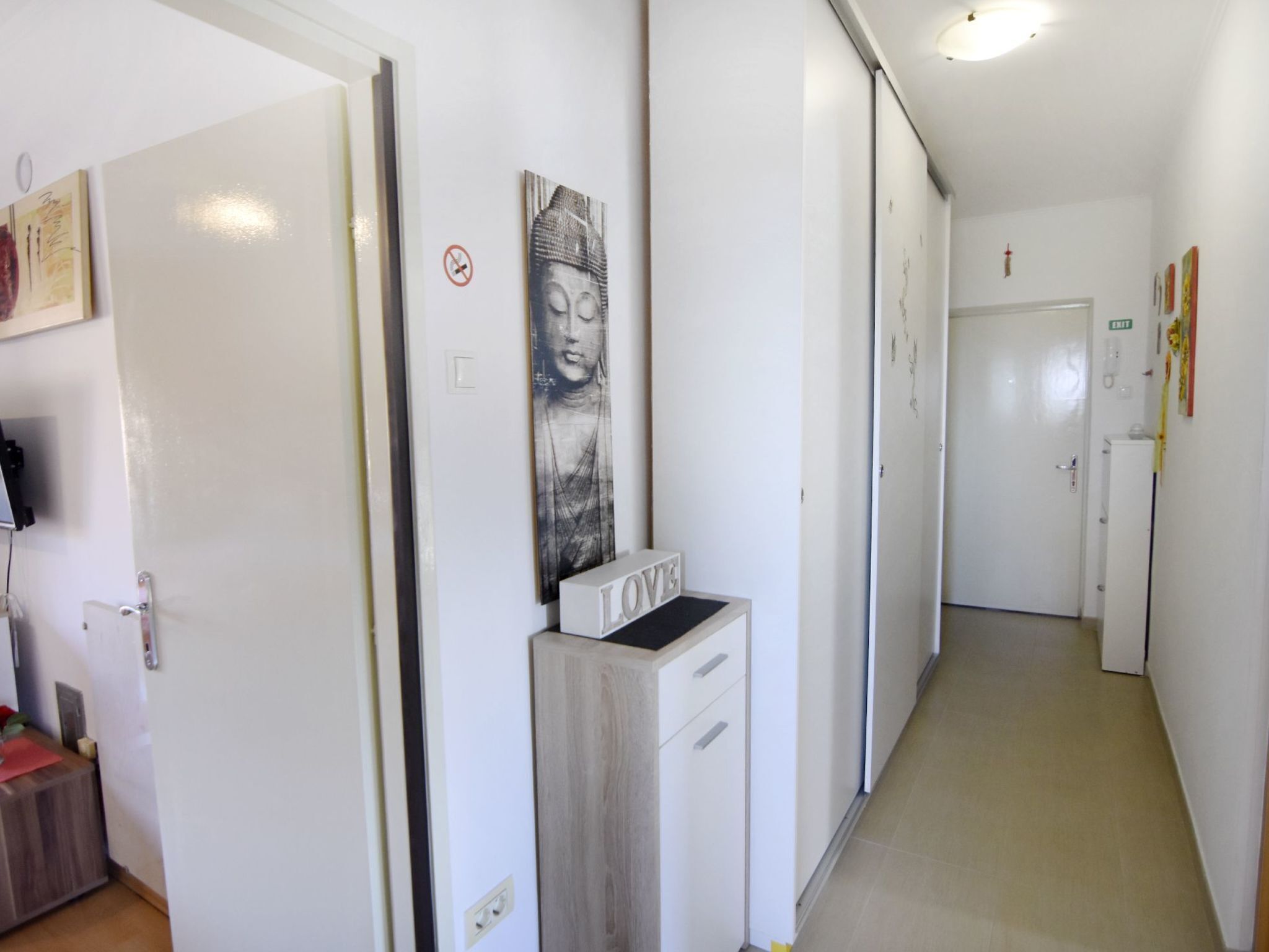 Ein Flur mit weißen Wänden, einer Tür mit einem 'Nichtraucher'-Schild, einem 'Love'-Schild auf einem Schrank und einem Buddha-Gemälde.