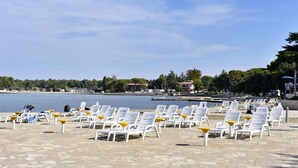 A waterfront area with rows of white lounge chairs and yellow tables, a stone-paved walkway, and a calm body of water in the background. - Martina B4 by Interhome (Umag)