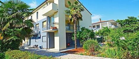 A modern two-story building with balconies, surrounded by lush greenery and palm trees.