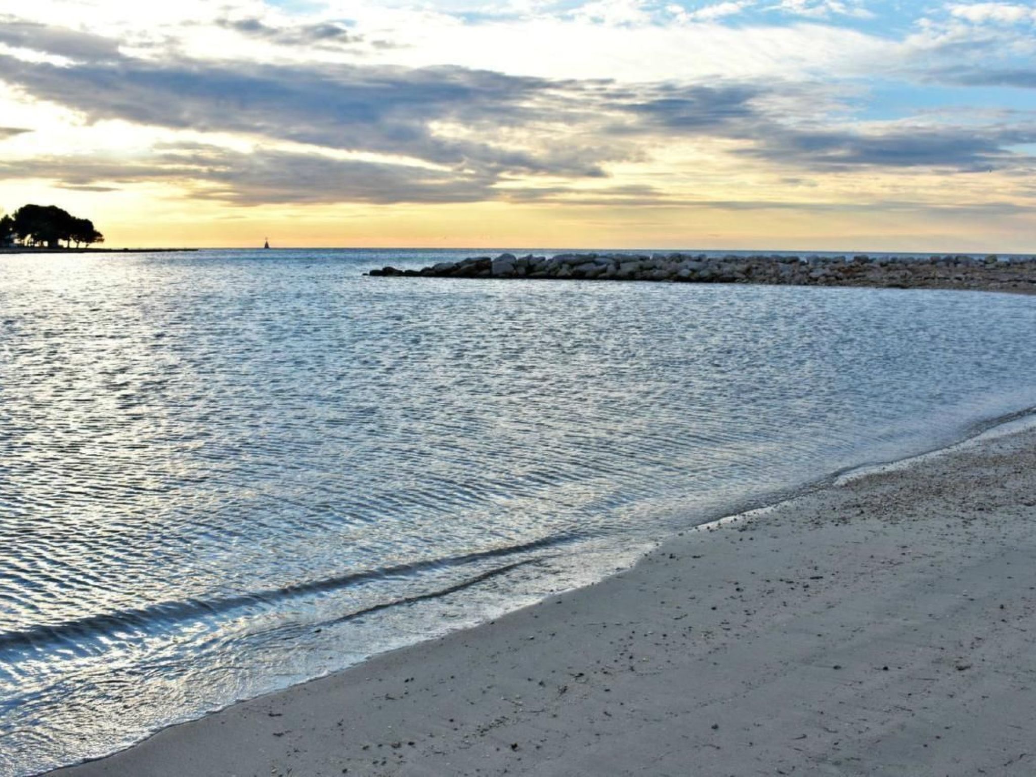 Una playa con agua tranquila, un rompeolas rocoso y un faro a lo lejos.