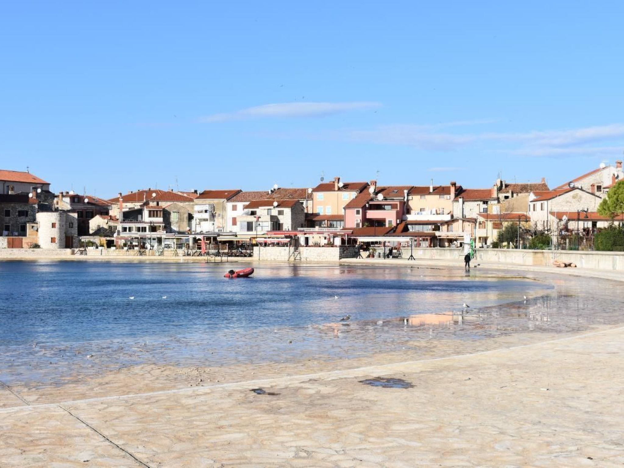 Una piazza sul lungomare, con edifici, una piccola barca e una persona che cammina.