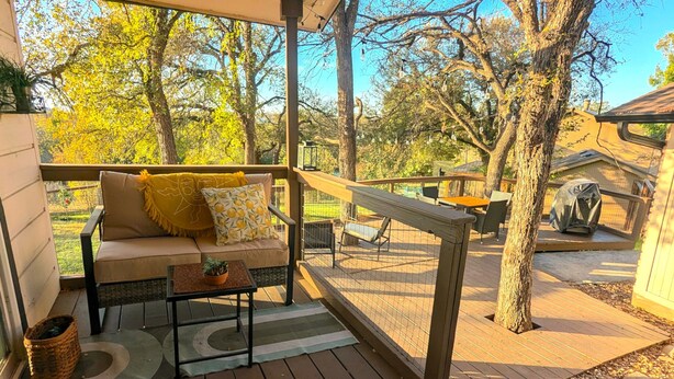 Un espacio al aire libre techado con sofá y mesa, rodeado de árboles y una terraza de madera.