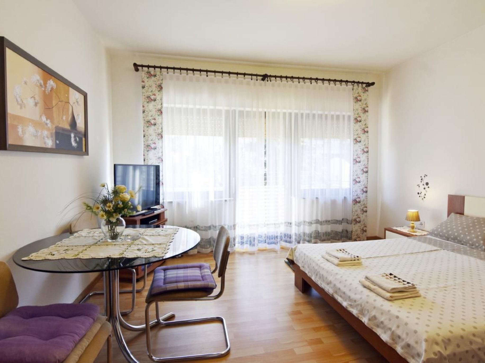 A hotel room with a bed, a TV, a dining table with chairs, and a window with curtains.
