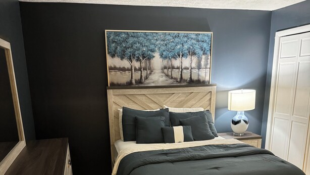 Un dormitorio con cama, mesita de noche, lámpara, cómoda y un cuadro en la pared.