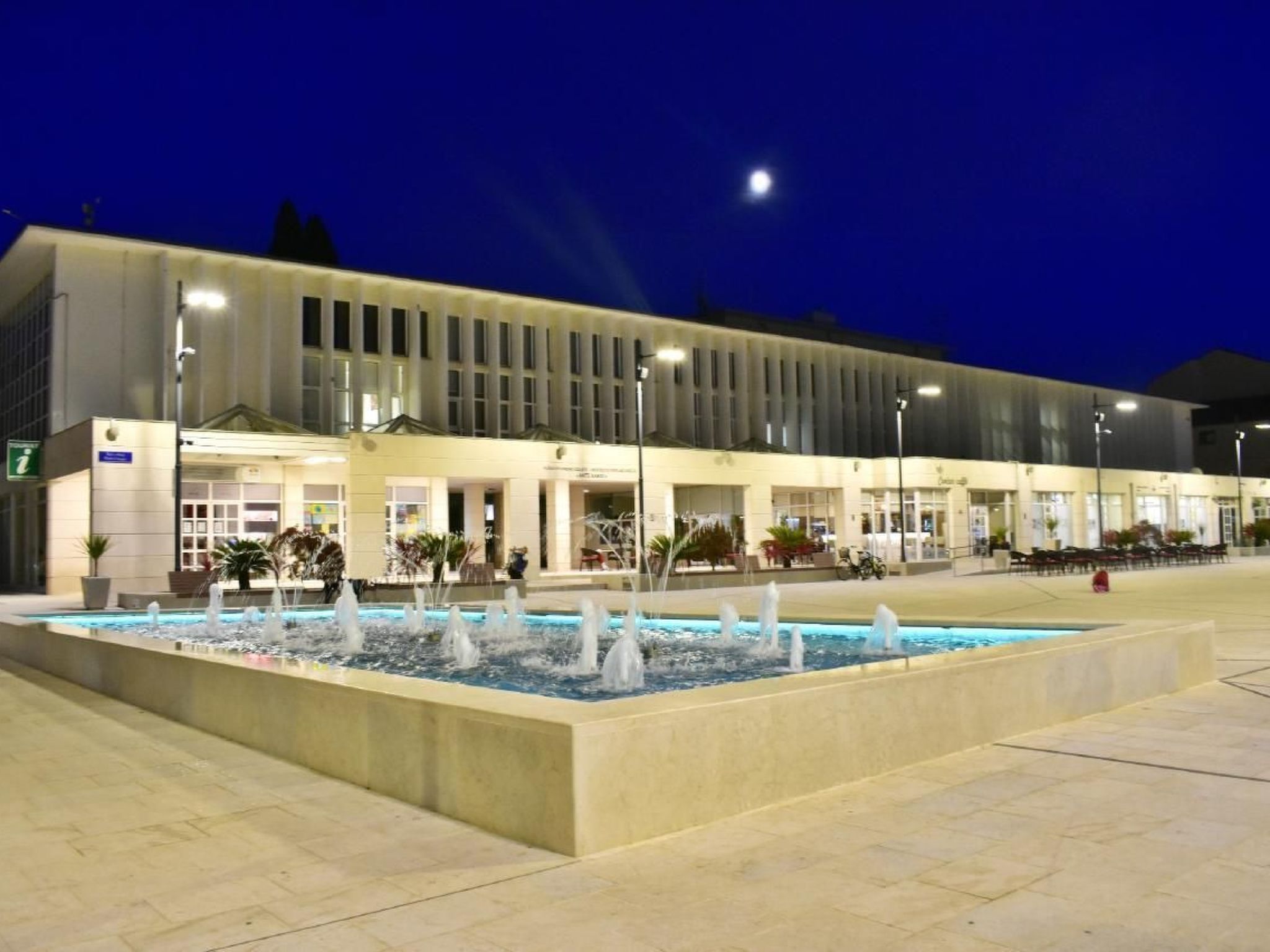 A modern building with a large outdoor fountain, a well-lit entrance, and a parking area.