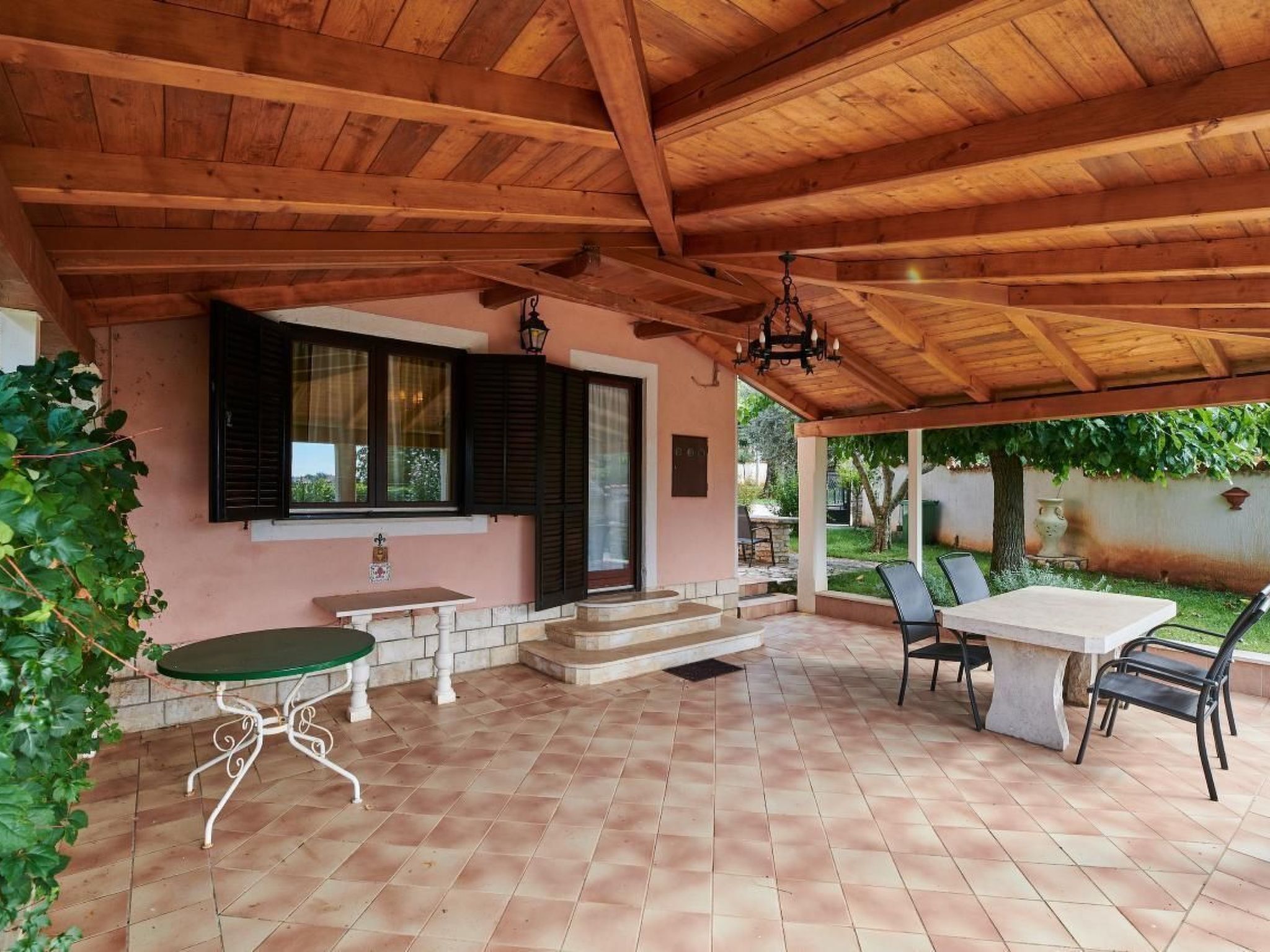 Eine überdachte Terrasse mit Holzboden, einem Tisch und Stühlen sowie einem kleinen Garten, der durch die Fenster sichtbar ist.