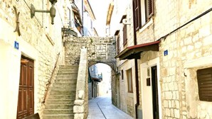 Una estrecha calle empedrada de piedra con una escalera que sube a un nivel superior, flanqueada por edificios con puertas y ventanas.