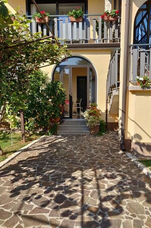 Un cortile lastricato in pietra con un edificio a due piani, un ingresso ad arco e piante in vaso.