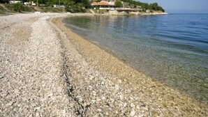 Ein Kiesstrand mit klarem Wasser, ein Gebäude mit rotem Dach und eine Hügelkette im Hintergrund.