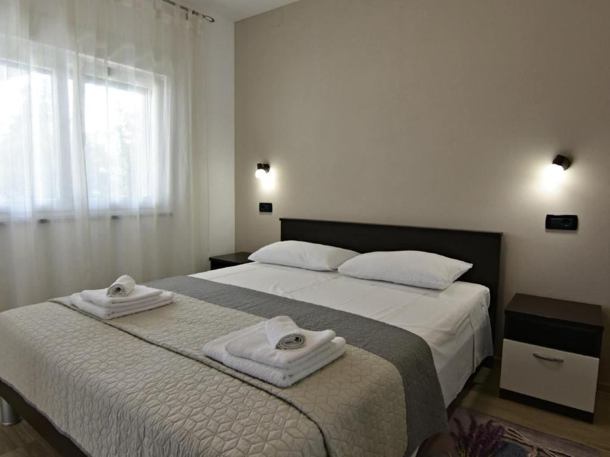 Cama bem arrumada com colcha cinza e branca, dois travesseiros brancos e duas toalhas enroladas. Há duas mesas de cabeceira com abajures e uma janela com cortinas translúcidas.