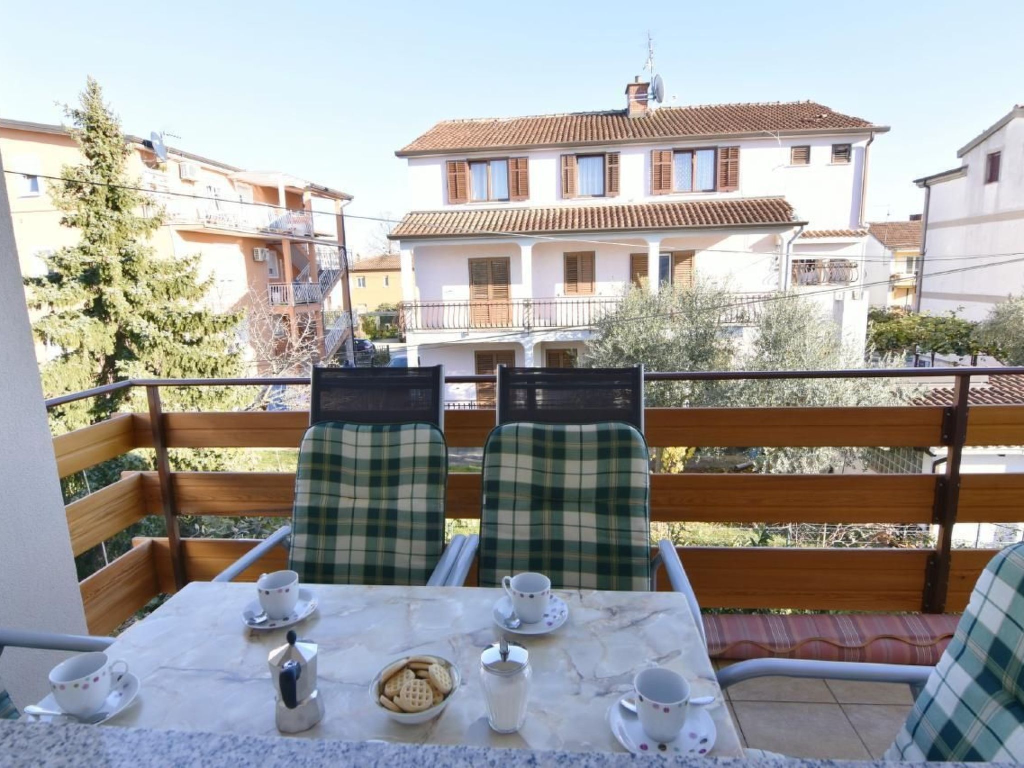 A balcony with two checkered chairs, a table with cups and a teapot, and a view of a building with a tiled roof.