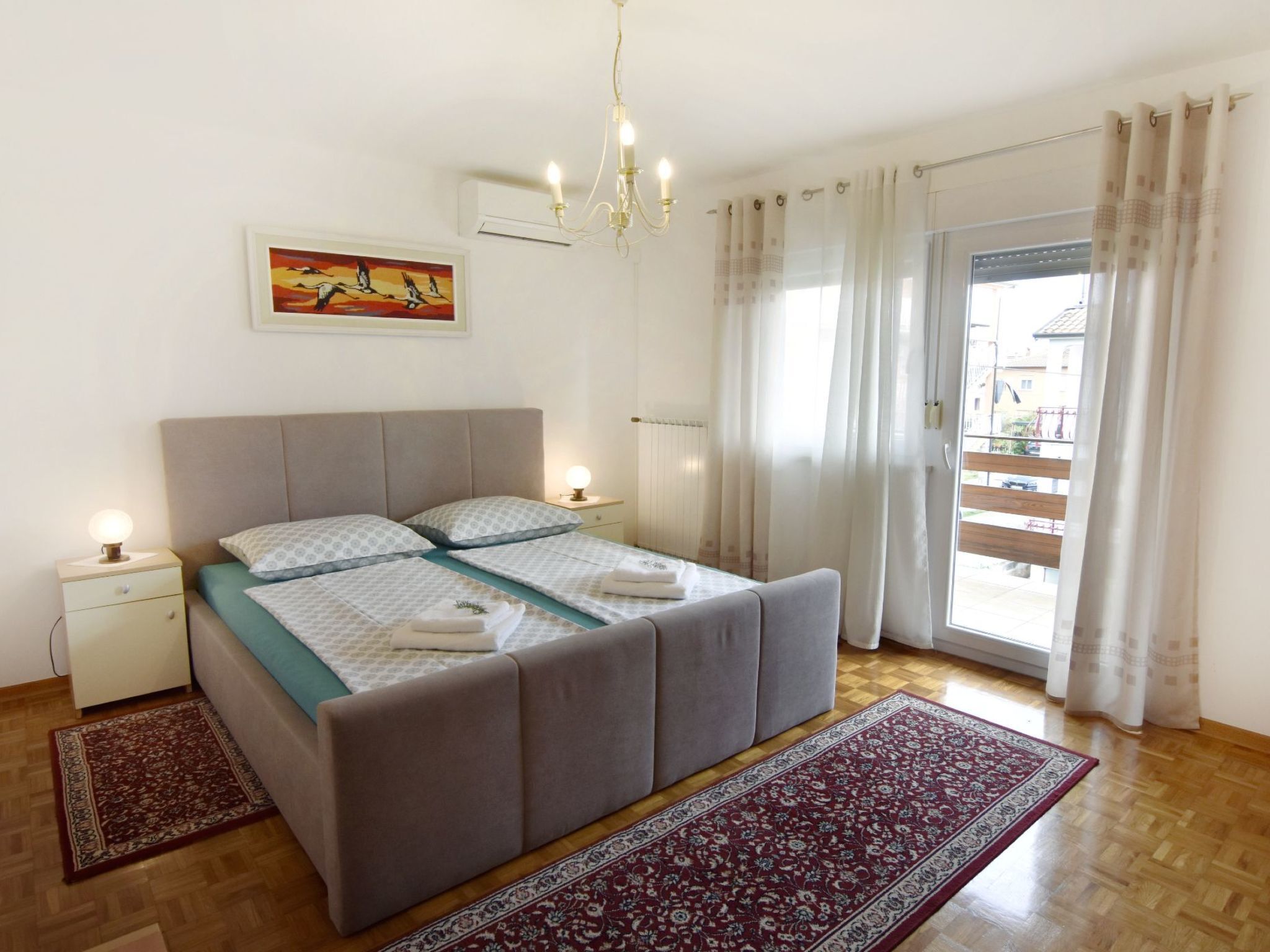 A bedroom with a bed, nightstands, a chandelier, a painting, and a view through a window.