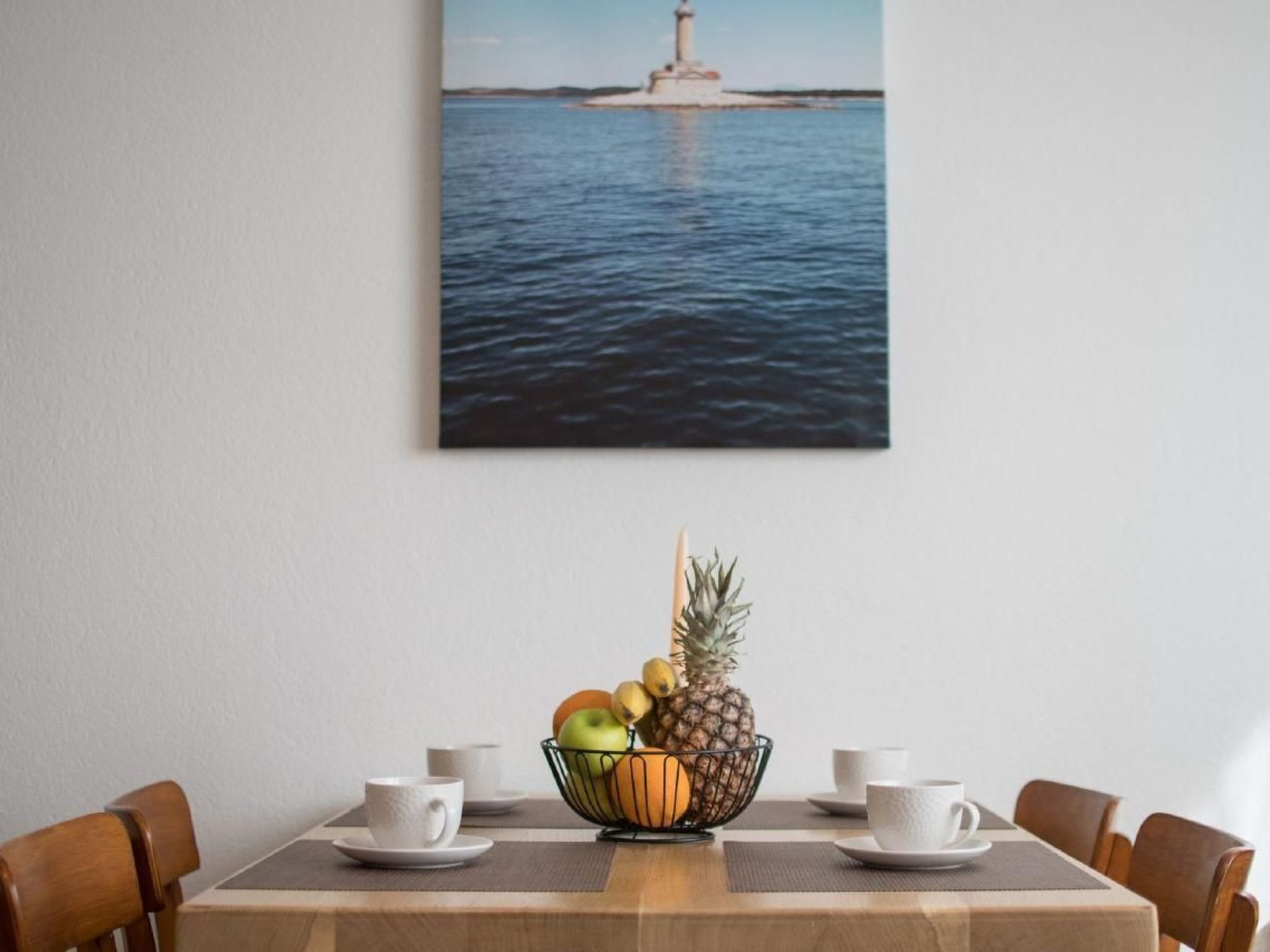 Una mesa de comedor con un frutero, tazas blancas y vistas a un faro sobre el agua.