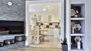 Een moderne woonkamer met een flatscreen televisie, een eethoek met een tafel en stoelen, en een keuken met witte kastjes en een zwarte spatwand.