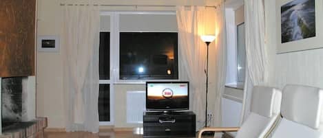 Una sala de estar con chimenea, televisor y un amplio ventanal con cortinas.