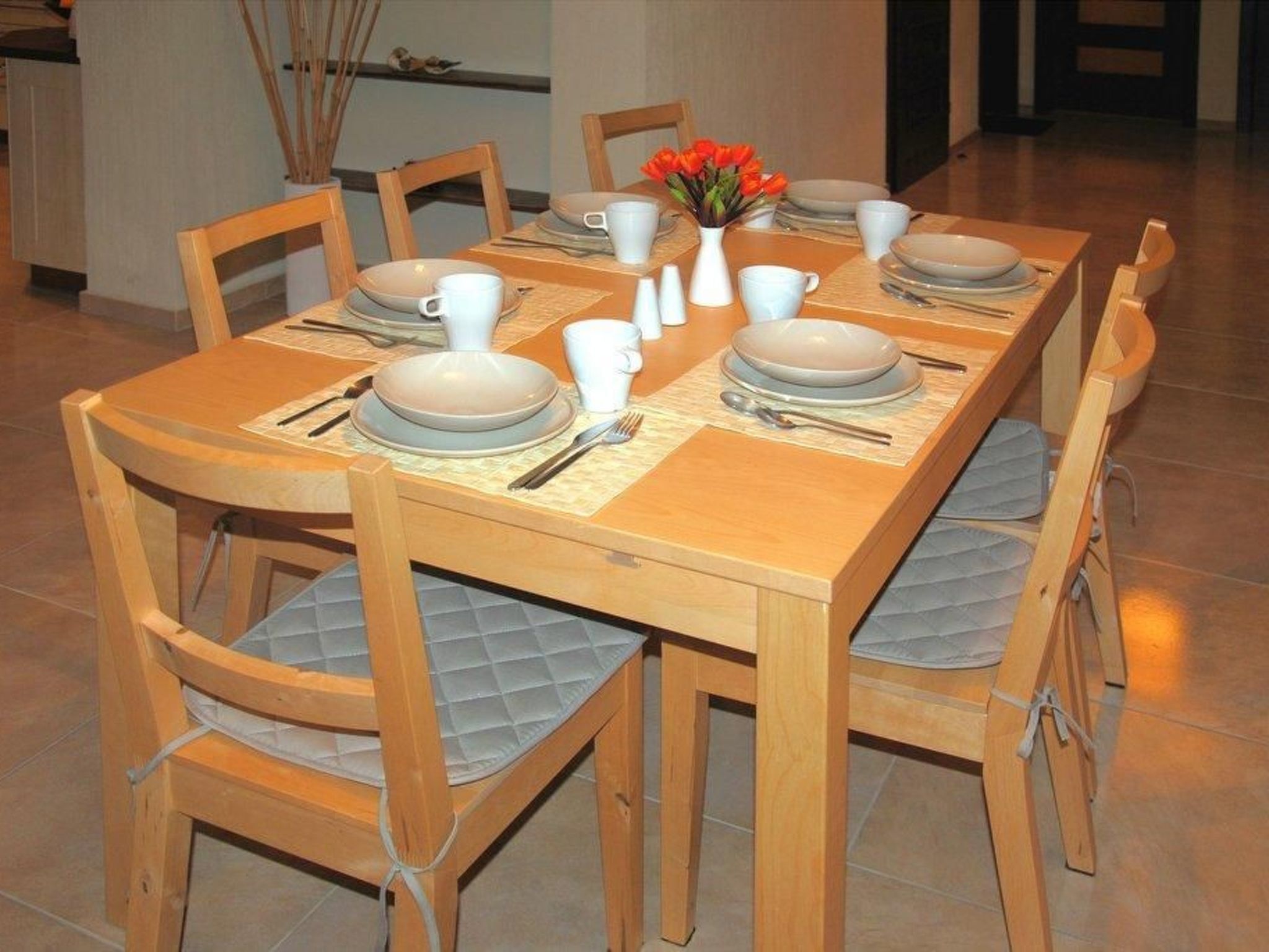 Ein Esstisch ist gedeckt mit Tellern, Tassen und Besteck, umgeben von Holzstühlen mit gepolsterten Sitzflächen.