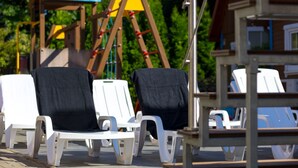 A row of white lounge chairs with black cushions, a wooden swing set, and a blue tarp in a sunny outdoor area. - "Apartment1" mit Gemeinschaftspool by Interhome (Rewal)