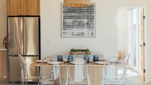 Eine moderne Küche mit einem Edelstahl-Kühlschrank, einem Holzesstisch mit klaren Stühlen und einer hängenden Lampenschirm aus gewebtem Material.