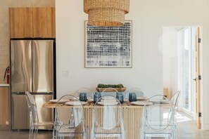 Eine moderne Küche mit einem Edelstahl-Kühlschrank, einem Holzesstisch mit klaren Stühlen und einer hängenden Lampenschirm aus gewebtem Material.