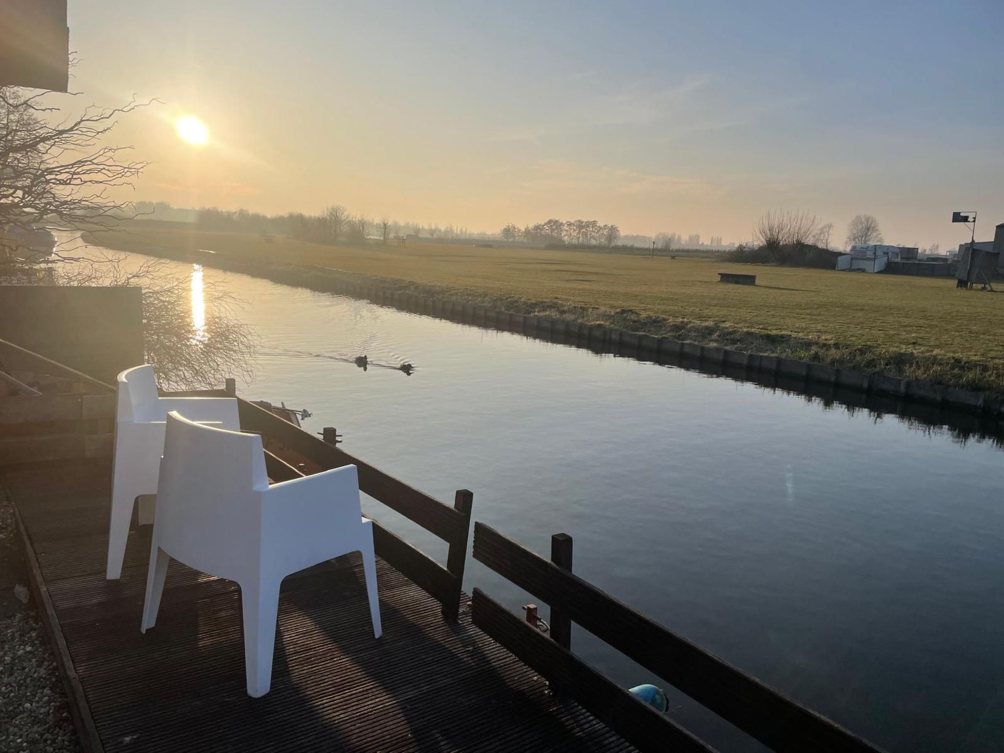 Blick auf einen Fluss mit einer Holzterrasse, weißen Stühlen und einem ruhigen Wasserspiegel, der die Sonne reflektiert.