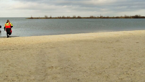 Una platja sorrenca amb una persona que camina per la riba portant un prim i un chaleco vermell i groc.