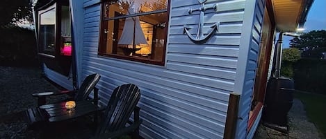 Una cabaña pequeña con una ventana que muestra un modelo de velero, un barco amarrado y una mesa con sillas afuera.