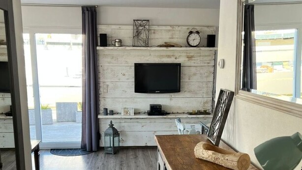 A modern living room with a wooden floor, a TV mounted on a white wall, and a large window.