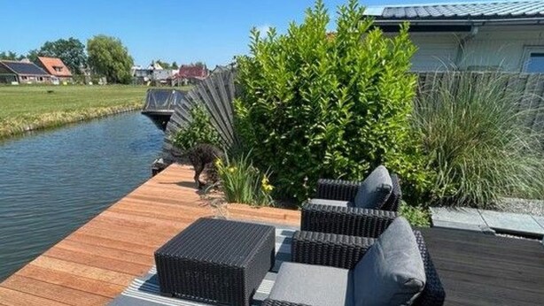 Una terraza de madera con muebles de exterior, un pequeño cuerpo de agua y follaje.