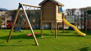 Een houten speelhuisje met een glijbaan en schommels in een gazon.