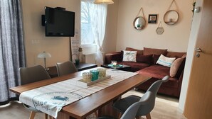 Een moderne woonkamer met een houten eettafel, een bank en een aan de muur bevestigde televisie.