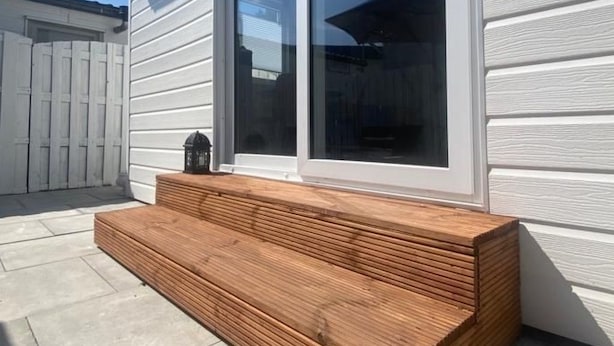 Un banc en bois se trouve devant une maison dont le revêtement extérieur est blanc et qui possède une grande porte-fenêtre coulissante.