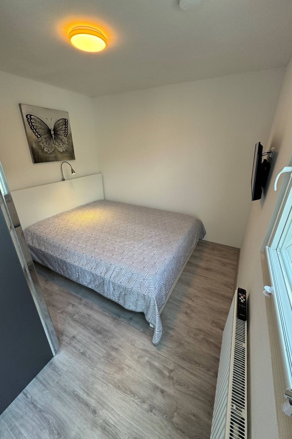 Une petite chambre à coucher comprenant un lit, un téléviseur fixé au mur, un radiateur et un luminaire au plafond.