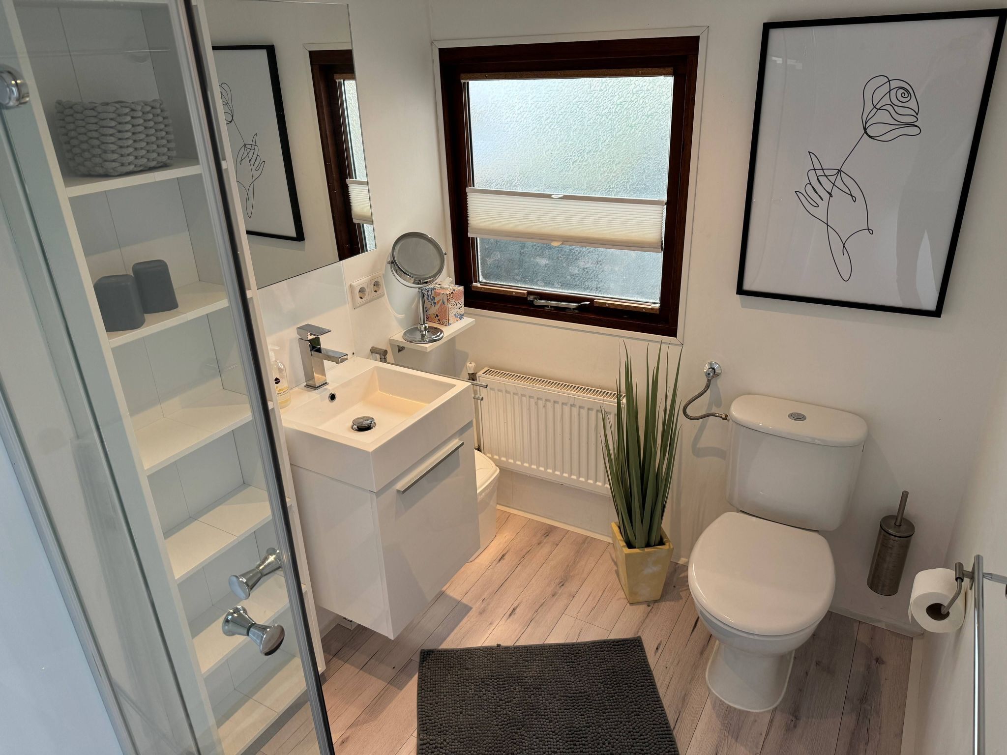 A modern bathroom with a white sink, mirror, toilet, and radiator. There is a window with a view, a potted plant, and a framed picture on the wall.