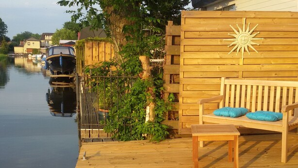 A wooden deck with a bench and a table, a wooden fence with a sun decoration, and a calm water body with boats. - Haus Jule by Interhome (Wervershoof)