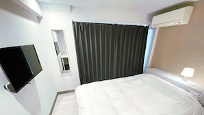 Hotellihuone, jossa on sänky, seinälle asennettu taulutelevisio, tummilla verhoilla varustettu ikkuna ja sängyn yläpuolella oleva ilmastointilaite.