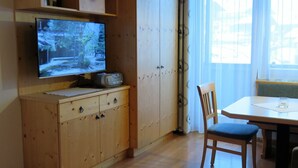 Ein Zimmer mit einem Holzkasten, einem Fernseher, einem Esstisch mit Stühlen und einem Fenster mit durchsichtigen Vorhängen.