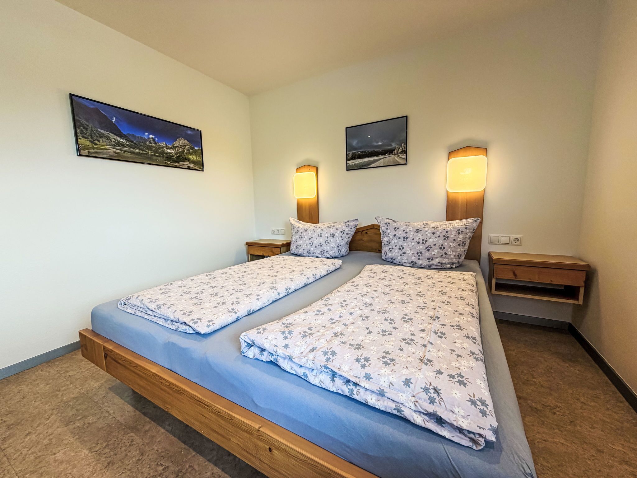 A double bed with floral bedding, two bedside tables, wall-mounted lamps, and two framed pictures.