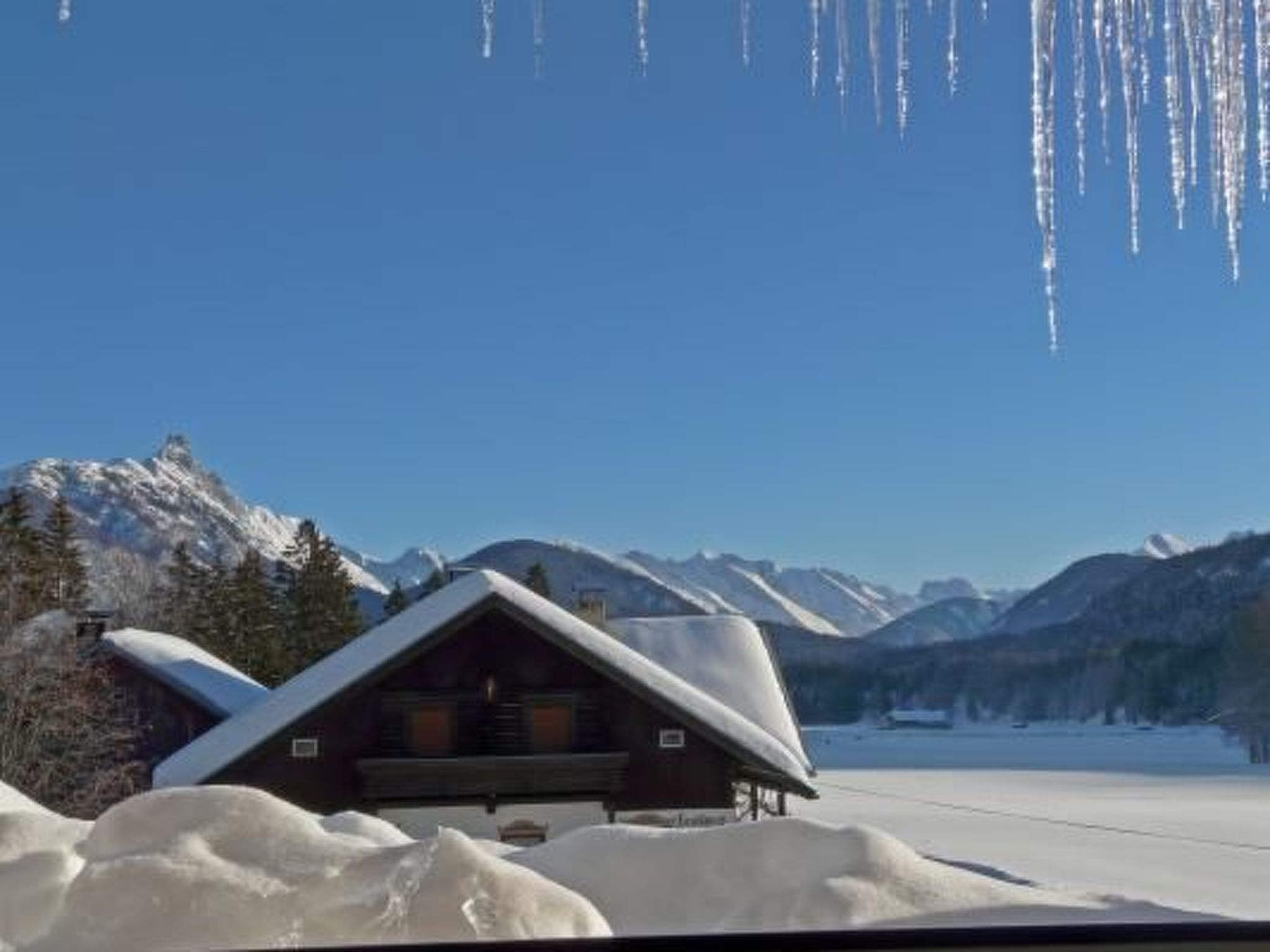 A cabin with a sloped roof, surrounded by snow-covered mountains and a clear sky.