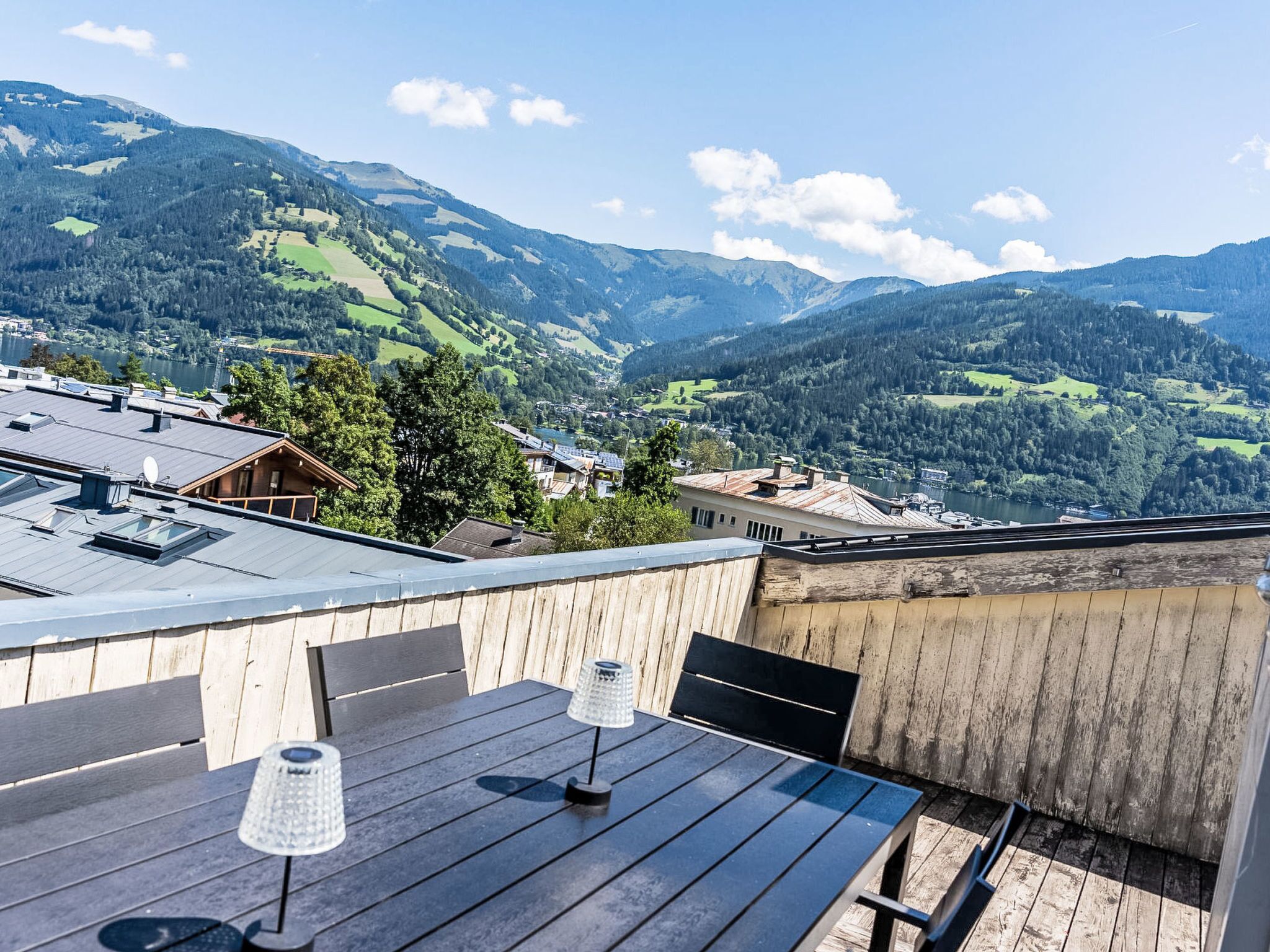 Eine Holzterrasse mit Tisch und Stühlen, mit Blick auf eine Berglandschaft mit See und Gebäuden.