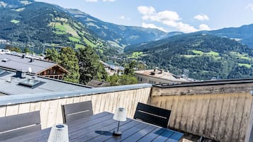 Eine Holzterrasse mit Tisch und Stühlen, mit Blick auf eine Berglandschaft mit See und Gebäuden.