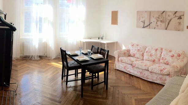 Un comedor con suelo de madera en patrón de espiga, una mesa de comedor dispuesta con platos y vasos, y un sofá floral.
