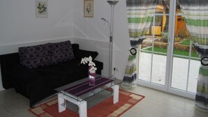 A living room with a black sofa, a glass coffee table, and a vase of flowers. There are two framed pictures on the wall, a floor lamp, and a sliding glass door with curtains. - Florian 1 in Bezirk 21-Floridsdorf by Interhome (Vienna)