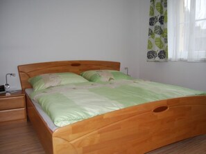 Ein Einzelbett mit Holzrahmen, einer grünen Bettdecke und passenden Kissen. Ein Nachttisch mit Lampe und Schublade ist vorhanden. Ein Fenster mit einem blattmusternden Vorhang ist sichtbar.